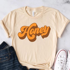 May include: A cream-colored short-sleeve t-shirt featuring the word Honey printed in a bold, retro-style orange and brown font across the chest. The shirt is paired with dark blue denim jeans and a pair of white canvas sneakers with laces.