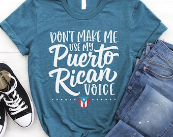 May include: A teal short-sleeve t-shirt featuring the white text "DON'T MAKE ME USE MY PUERTO RICAN VOICE" and a small Puerto Rican flag icon. The shirt is displayed alongside a pair of black canvas sneakers and blue denim jeans.