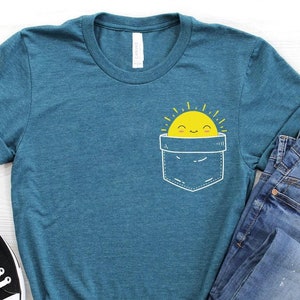 May include: Teal t-shirt with a pocket design featuring a smiling yellow sun. The shirt is paired with blue jeans and black canvas sneakers. The design is simple and cheerful, perfect for casual wear.