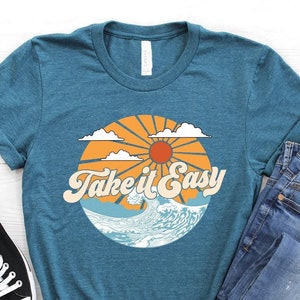 May include: Teal t-shirt with a retro graphic that says "Take it Easy". The graphic features a sun, clouds, and ocean waves. The shirt is paired with blue jeans and black canvas sneakers.