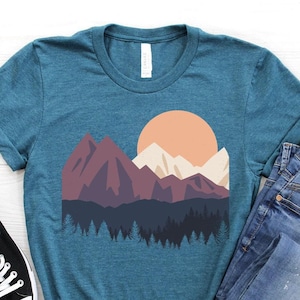 May include: Teal t-shirt featuring a mountain landscape graphic with a sunset. The design includes purple, white, and dark green elements. The shirt is paired with blue jeans and black canvas sneakers.