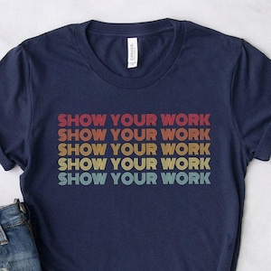 May include: Navy blue t-shirt with the text "SHOW YOUR WORK" in a retro, stacked design. The text is in red, orange, yellow, and green. The shirt is paired with denim shorts.