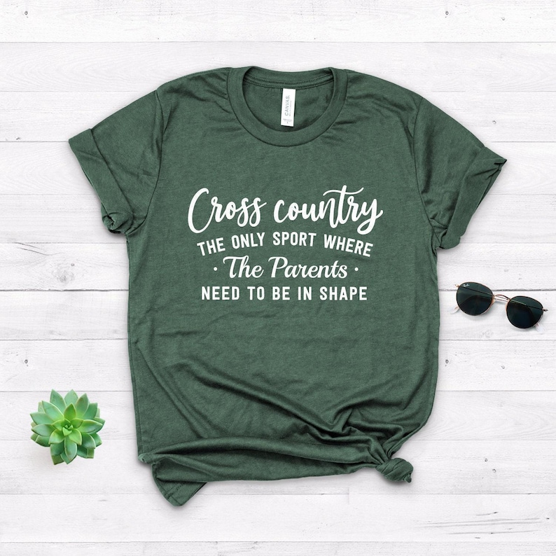 An olive green t-shirt with white lettering displaying Cross country THE ONLY SPORT WHERE The Parents NEED TO BE IN SHAPE is shown on a white wooden surface with sunglasses and a green succulent.