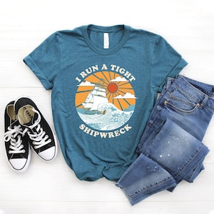 Könnte beinhalten: Ein türkisfarbenes T-Shirt mit einer Grafik im Vintage-Stil eines Schiffes, das in eine Welle fährt, mit dem Text "I run a tight shipwreck".
