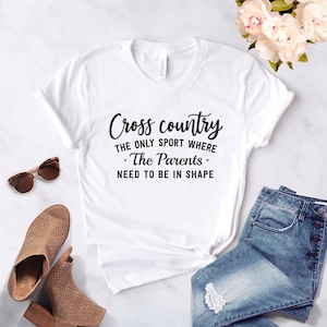 A white t-shirt printed with the black text Cross country THE ONLY SPORT WHERE The Parents NEED TO BE IN SHAPE is laid flat on a white marble surface next to denim shorts, sunglasses, brown sandals, and pink flowers.