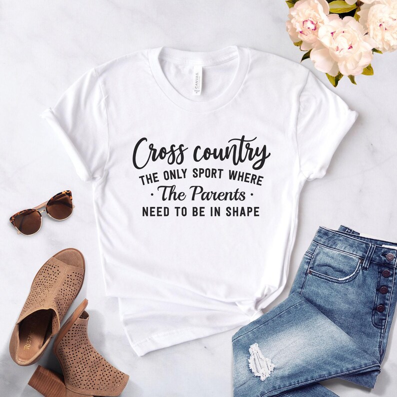 A white t-shirt printed with the black text Cross country THE ONLY SPORT WHERE The Parents NEED TO BE IN SHAPE is laid flat on a white marble surface next to denim shorts, sunglasses, brown sandals, and pink flowers.