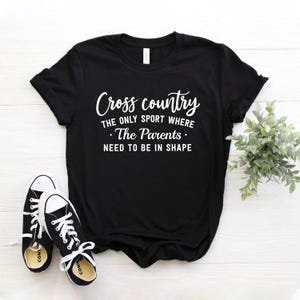 A black t-shirt featuring white lettering that says Cross country THE ONLY SPORT WHERE The Parents NEED TO BE IN SHAPE rests on a white wooden tabletop beside black-and-white sneakers and a small green potted plant.