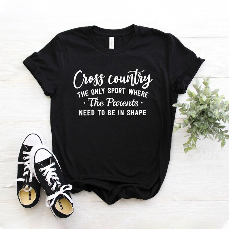 A black t-shirt featuring white lettering that says Cross country THE ONLY SPORT WHERE The Parents NEED TO BE IN SHAPE rests on a white wooden tabletop beside black-and-white sneakers and a small green potted plant.