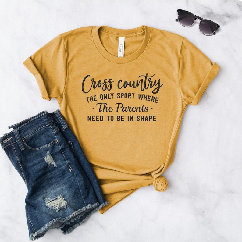 A mustard yellow t-shirt printed with black text reading Cross country THE ONLY SPORT WHERE The Parents NEED TO BE IN SHAPE is displayed on a white surface alongside folded blue denim shorts and sunglasses.