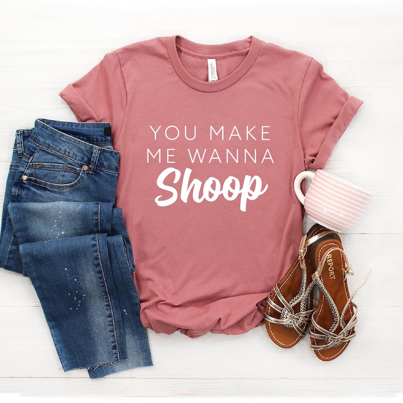 May include: A dusty rose t-shirt with the text "You make me wanna Shoop" in white lettering. The shirt is paired with blue jeans and brown sandals.