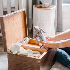 May include: A rectangular wooden storage chest with a hinged lid, featuring a dark walnut exterior and a light-colored interior with a removable divider. Inside the chest, a white plush sheep toy rests alongside a grey fabric rabbit toy.