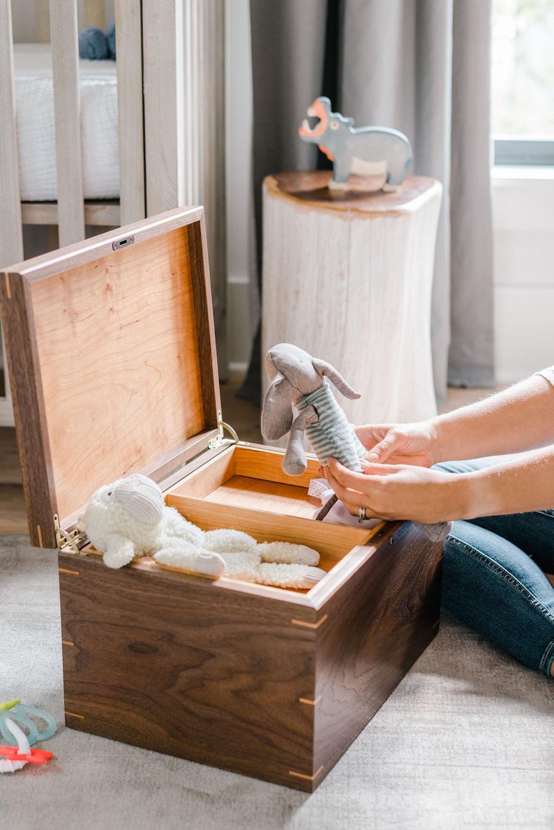 May include: A rectangular wooden storage chest with a hinged lid, featuring a dark walnut exterior and a light-colored interior with a removable divider. Inside the chest, a white plush sheep toy rests alongside a grey fabric rabbit toy.