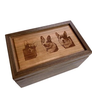 May include: A rectangular wooden box featuring a dark brown frame and a lighter brown lid. The lid is engraved with three detailed portraits of Corgi dogs, each showing a distinct facial expression and posture, centred on the smooth wood surface.