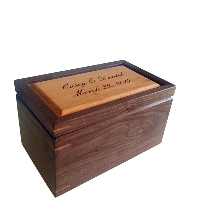 May include: A rectangular wooden box featuring a dark brown walnut body and a lighter-toned wood lid. The lid is engraved with the text Casey & Daniel March 23, 2019 in a cursive script. The box displays visible joinery details at the corners.