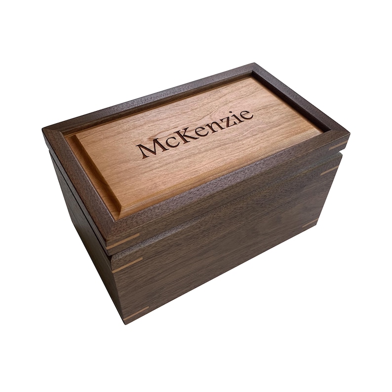 May include: A rectangular wooden box featuring a dark brown walnut exterior and a lighter brown wood lid. The name McKenzie is engraved in a serif font on the centre of the lid. The box displays visible joinery details with light wood accents.