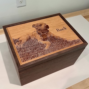 May include: A rectangular wooden box with a dark brown frame and a light wood lid. The lid features a laser-engraved image of a dog lying in grass and the name Rock engraved in a dark font. The box has visible joinery details on the corners.