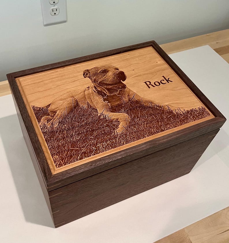 May include: A rectangular wooden box with a dark brown frame and a light wood lid. The lid features a laser-engraved image of a dog lying in grass and the name Rock engraved in a dark font. The box has visible joinery details on the corners.