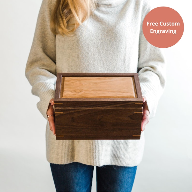 Woman holding a handcrafted walnut keepsake box with a cherry wood lid and corner spline joinery, showcasing a modern heirloom design with free custom engraving included.