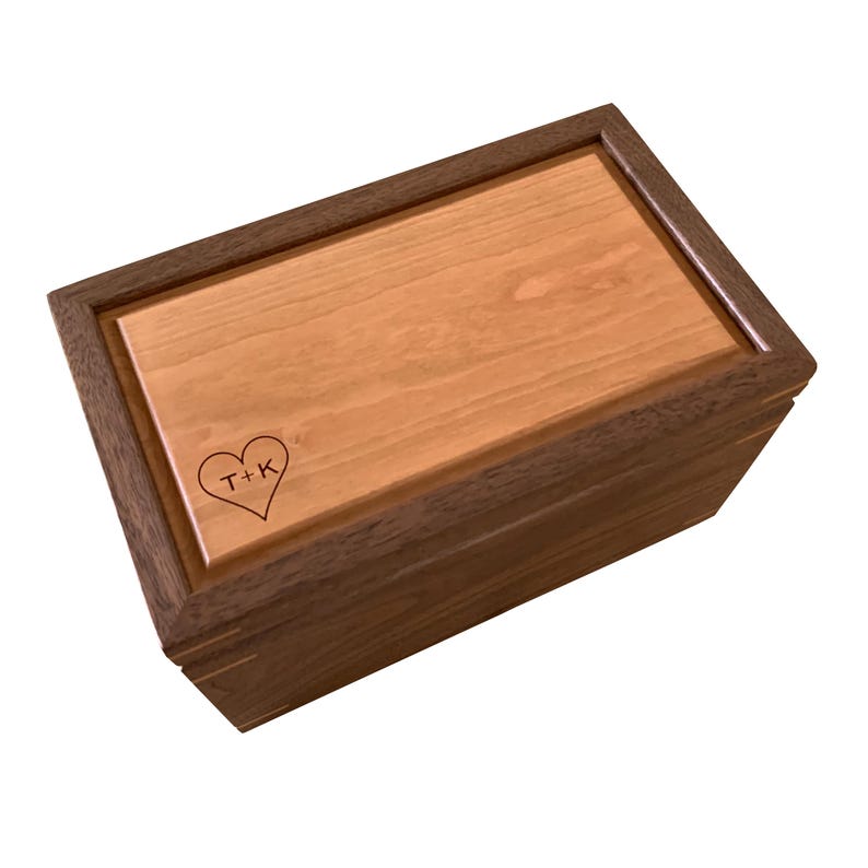 May include: A rectangular wooden box featuring a dark brown frame and a lighter tan wood lid. The lid is engraved with a heart shape containing the letters T+K. The box has a smooth finish and visible wood grain patterns on the top and sides.
