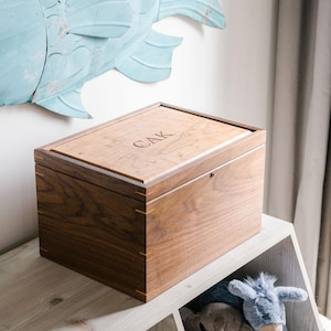 May include: A rectangular wooden keepsake box crafted from dark walnut wood with light wood corner splines. The lid features a lighter wood inlay with the engraved letters CAK centered on the top surface. The box has a smooth finish.