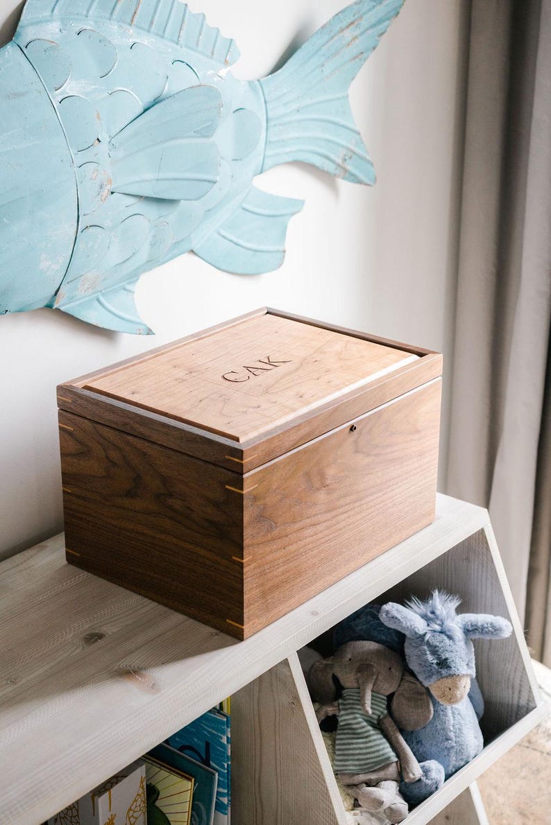 May include: A rectangular wooden keepsake box crafted from dark walnut wood with light wood corner splines. The lid features a lighter wood inlay with the engraved letters CAK centered on the top surface. The box has a smooth finish.