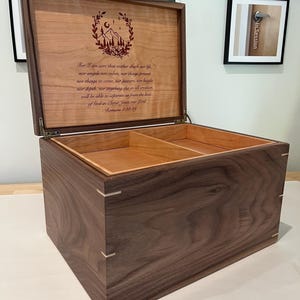 May include: A rectangular wooden keepsake box with a dark walnut exterior and light wood corner splines. The lid is open, revealing a light-colored interior with a divider and a laser-engraved mountain illustration and the text of Romans 8:38-39.