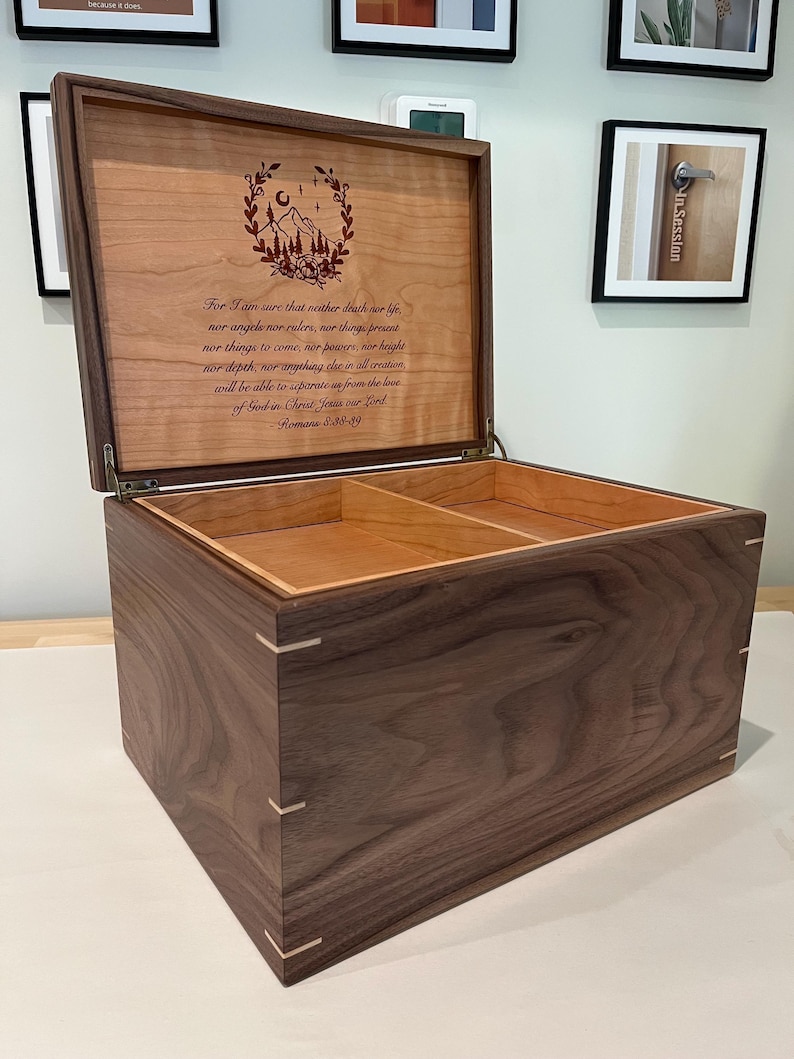 May include: A rectangular wooden keepsake box with a dark walnut exterior and light wood corner splines. The lid is open, revealing a light-colored interior with a divider and a laser-engraved mountain illustration and the text of Romans 8:38-39.