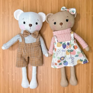 Puede incluir: Dos osos de peluche, uno con un atuendo de cuadros marrones y blancos y una corbata de moño, y el otro con un suéter rosa y una falda floral con fresas.