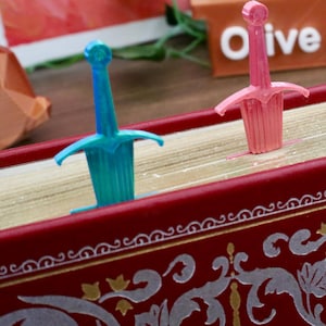 May include: Two plastic sword-shaped bookmarks, one pink and one blue, are placed on a red book with gold accents and a decorative pattern. The book is closed and the bookmarks are sticking out of the top.