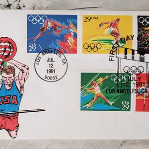 May include: A commemorative envelope with a stamp featuring a pole vaulter holding an American flag. The envelope has a postmark from Los Angeles, California, July 12, 1991, and four other stamps depicting Olympic events.