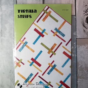 Könnte beinhalten: Ein Quilt-Anleitungsheft mit dem Titel Tortilla Strips von Atkinson Designs. Das Cover zeigt einen weißen Hintergrund mit einem verstreuten Muster aus sich kreuzenden rechteckigen Streifen in Rot, Blau, Orange und Grün. Der Code ATK-168 ist sichtbar.