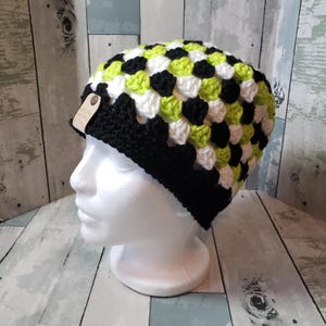 May include: A handmade crochet beanie in a granny square pattern. The hat features a black brim and a top section with a pattern of white, black, and neon green squares. A small tag is attached to the brim.