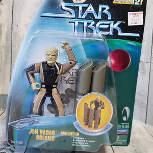 May include: A Star Trek Jem'Hadar Soldier action figure in its original packaging. The figure is tan, black, and brown, with articulated arms. The packaging includes the Star Trek logo and the text "Warp Factor Series 2".