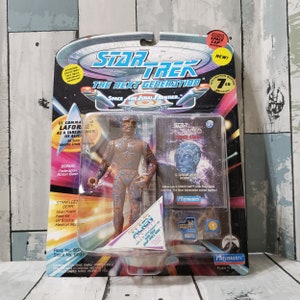 May include: A Star Trek: The Next Generation action figure of Lt. Commander La Forge as a Tarchwen alien. The figure is blue and gray with a translucent blue body. The figure is packaged in a plastic blister pack with a blue and white background. The packaging includes the text "Star Trek: The Next Generation" and "Space: The Final Frontier."