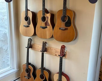 Może przedstawiać: Sześć gitar akustycznych jest zamontowanych na dwóch poziomych drewnianych uchwytach ściennych. Kolekcja obejmuje różne instrumenty w odcieniach drewna o odmiennych kształtach korpusów, maskownicach i główkach, ułożone w dwóch rzędach po trzy gitary.