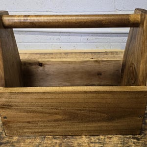 May include: A handcrafted wooden tool caddy with a handle. The caddy is made of light brown wood with a darker brown handle. The sides are angled, and the caddy is designed to hold tools or other small items. The wood grain is visible.
