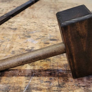 May include: A wooden mallet with a dark brown finish and a natural wood handle. The mallet is lying on a wooden surface.