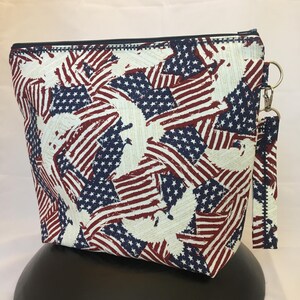 May include: A zippered pouch with a white background and a pattern of red, white, and blue American flags with distressed edges. The pouch has a black zipper and a black zipper pull with a silver ring.