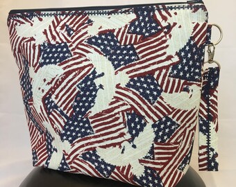 May include: A zippered pouch with a white background and a pattern of red, white, and blue American flags with distressed edges. The pouch has a black zipper and a black zipper pull with a silver ring.