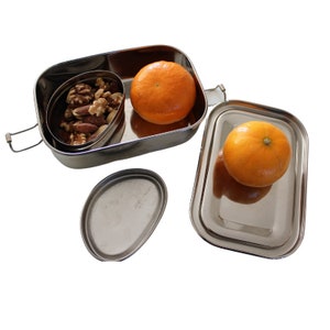Puede incluir: Una fiambrera de acero inoxidable con dos compartimentos. El compartimento superior contiene una mezcla de nueces y el compartimento inferior contiene dos naranjas enteras.