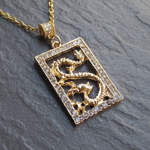 Puede incluir: Un collar de oro con un colgante que presenta un diseño de dragón rodeado de diamantes de imitación transparentes. El colgante tiene forma rectangular.