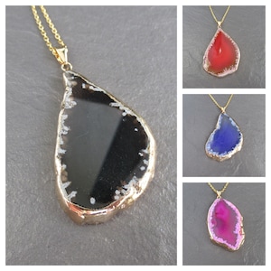 May include: A gold chain necklace with a pendant made of a large, teardrop-shaped slice of agate. The agate is translucent and has a black, white, and grey pattern. The necklace is shown in three different colours: red, blue, and pink.