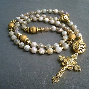 May include: A gold-toned rosary with a cross pendant and a large, ornate bead. The rosary is made with light grey and white beads.