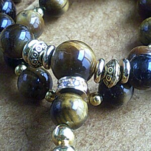 K&ouml;nnte beinhalten: Ein braunes und goldenes Perlenarmband mit silbernen Akzenten. Die Perlen sind aus Tigerauge und haben eine nat&uuml;rliche, polierte Oberfl&auml;che.
