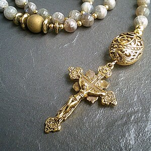May include: A gold-toned rosary necklace with a detailed cross pendant and a round, ornate bead. The necklace is made with grey and white stones.