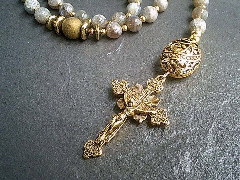 May include: A gold-toned rosary necklace with a detailed cross pendant and a round, ornate bead. The necklace is made with grey and white stones.