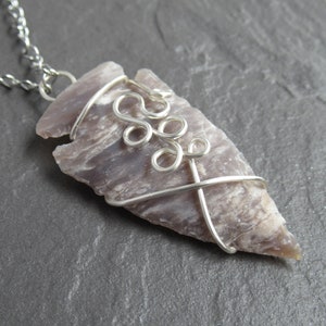 Puede incluir: Un collar de cadena de plata con un colgante hecho de una punta de flecha gris y blanca envuelta en alambre de plata. El alambre está retorcido en un diseño decorativo.
