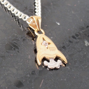 Puede incluir: Un collar con un colgante de nave espacial de oro y plata, con un strass transparente en el centro. El colgante está en una cadena de plata.