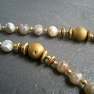 May include: A necklace made with grey and gold beads. The necklace features a mix of round and flat beads, with some of the round beads having a metallic finish.