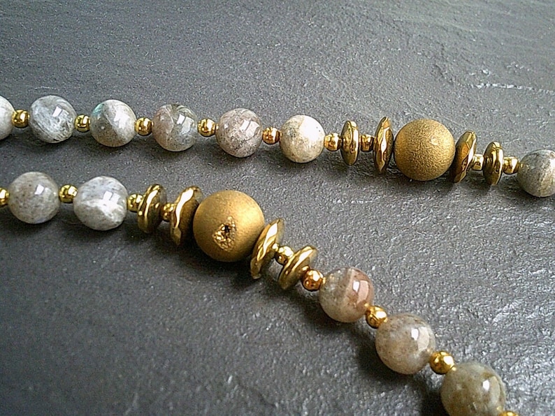 May include: A necklace made with grey and gold beads. The necklace features a mix of round and flat beads, with some of the round beads having a metallic finish.